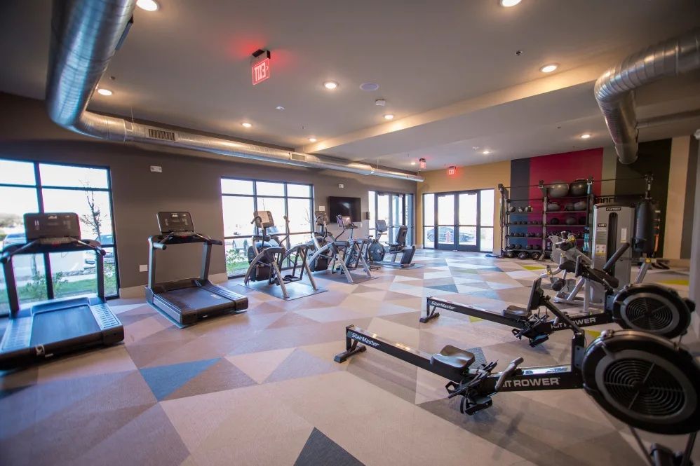 Gym with exercise equipment like treadmills, rowers, and weights; neutral-toned walls, and large windows.