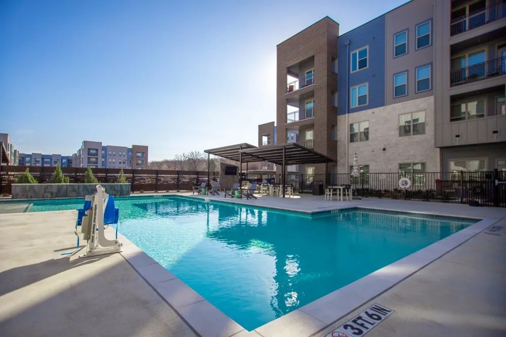 Pool with buildings in background. Clear water, blue sky, concrete deck, shade structure.