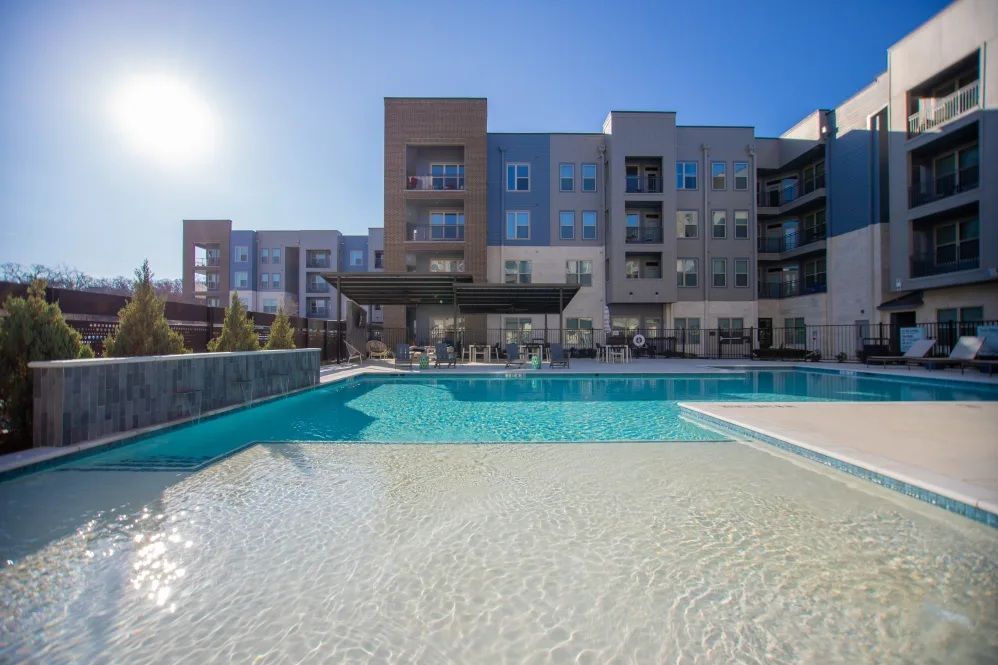 Pool with a white shallow area, in front of a modern apartment building under a sunny sky.