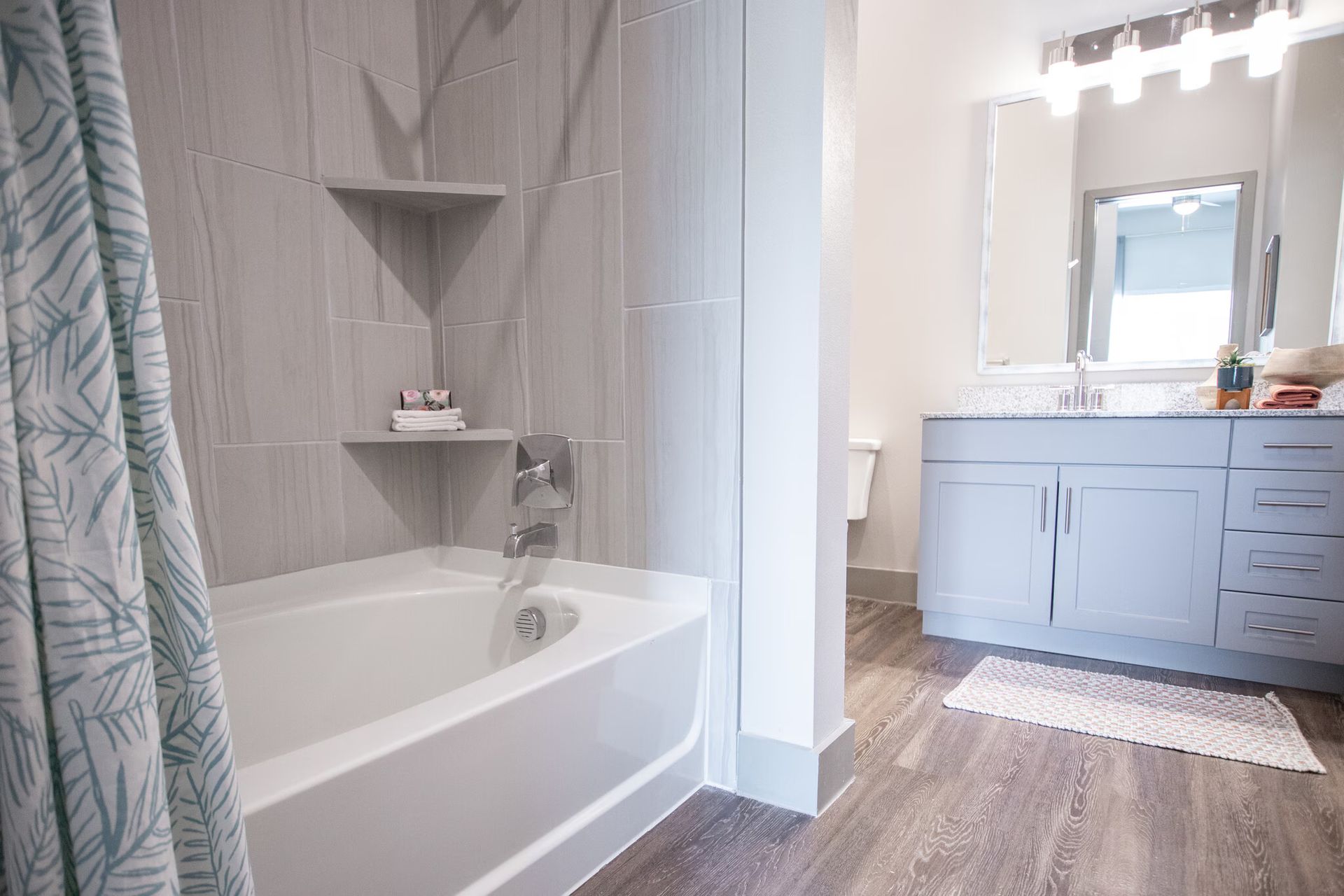 Bathroom with white bathtub, gray tile shower, and vanity with sink. Wooden floor.