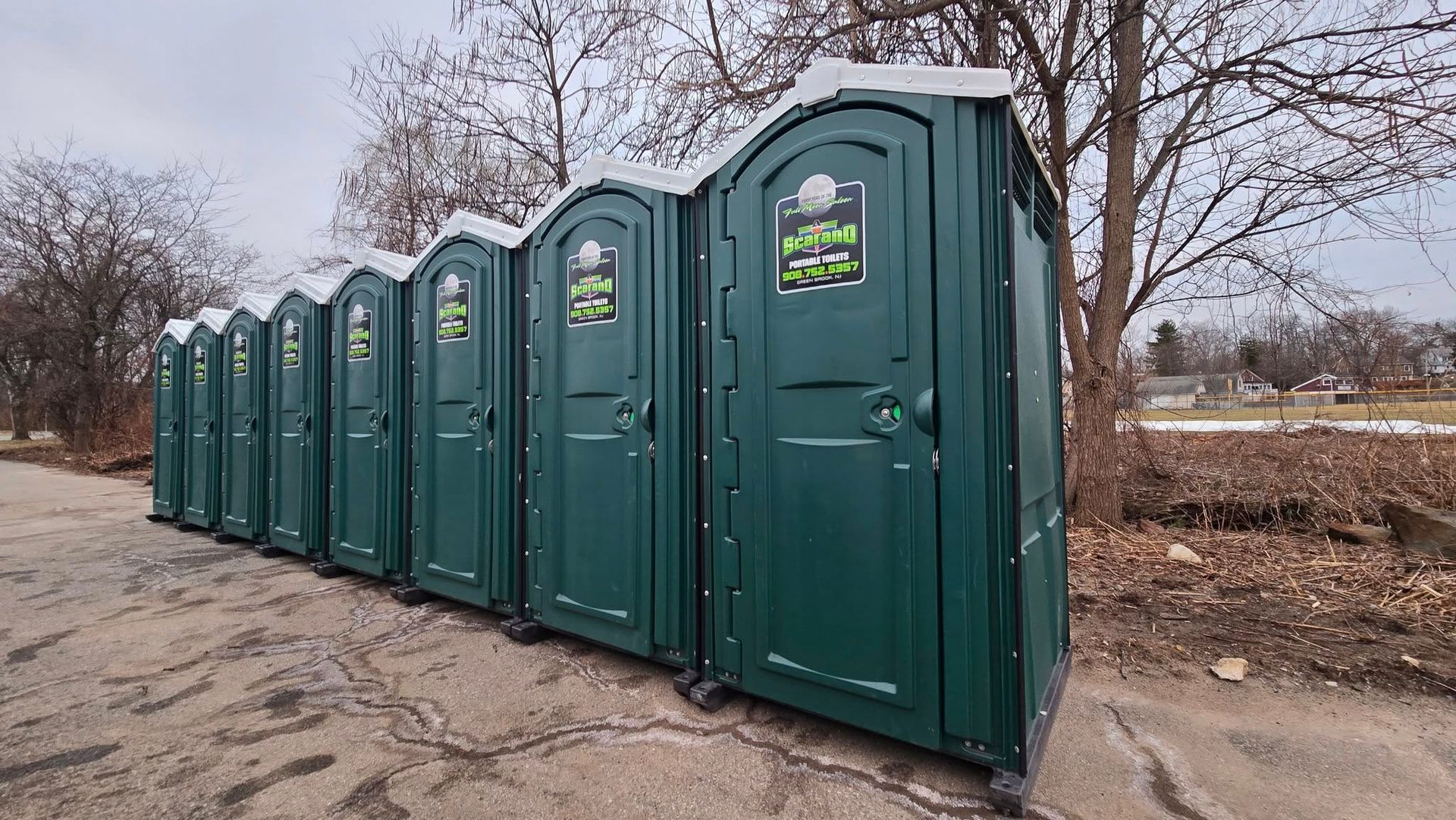 A row of eight dark green portable toilets in an outdoor setting on a gray day.