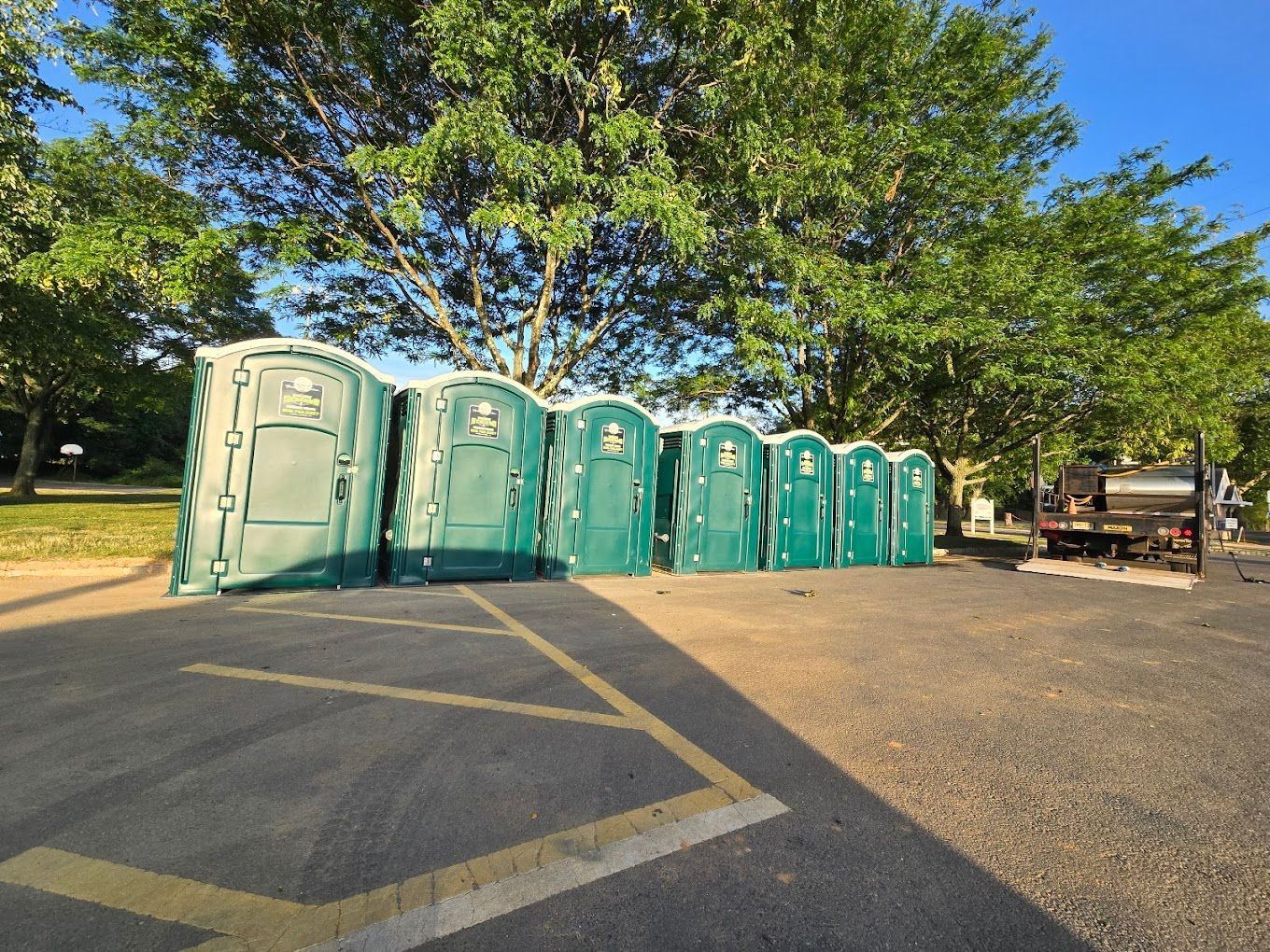 Row of green portable toilets in a parking lot, near trees.