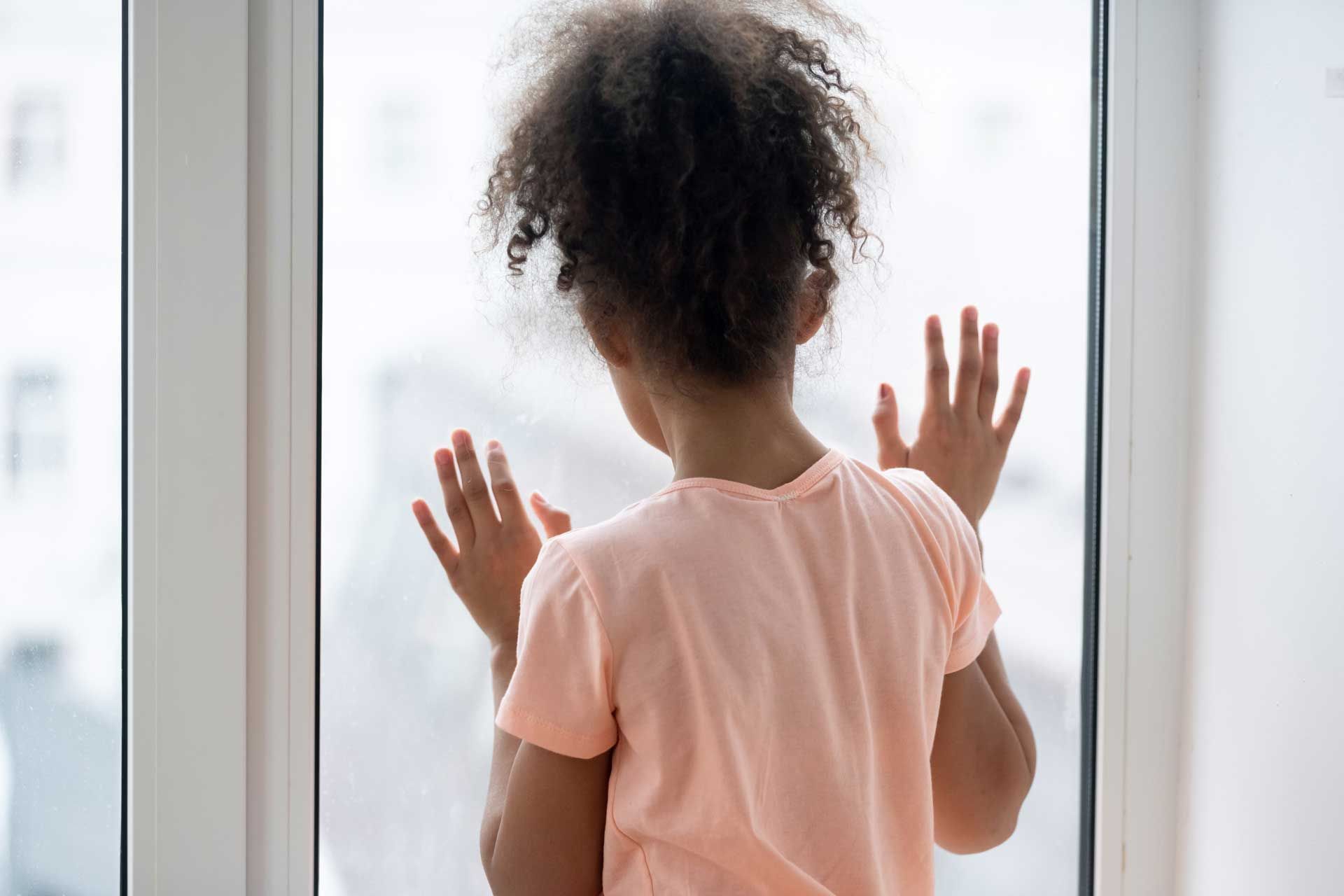 A little girl is looking out of a window with her hands on the glass.