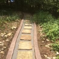 Narrow stone stepping path through a wooded garden, bordered by low edging and greenery