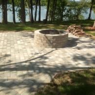 Stone fire pit on a paved patio beside a lake, with stacked bricks and trees in the background
