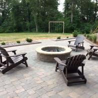 Backyard patio with a circular fire pit, Adirondack chairs, and a grassy yard with trees.