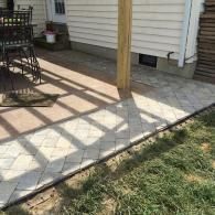Covered patio with stone pavers, wooden post, and shadows beside a house and grassy yard