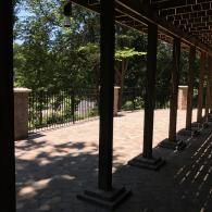 Shaded porch with black columns overlooking a sunlit walkway and trees beyond