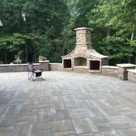 Stone outdoor patio with built-in brick fireplace, chairs, and wooded background