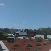 White pickup truck parked beside a landscaped red mulch area with flowers under a clear blue sky.
