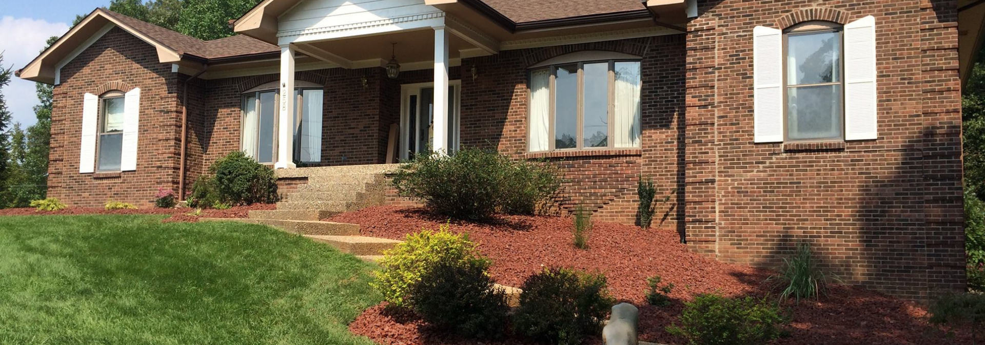 Brick house with white shutters, front porch, and landscaped lawn on a sunny day
