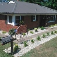 Front yard garden with a brick house, black mailbox, gravel path, and a decorative wagon wheel planter.