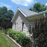 Side of a stone house with shrubs, green lawn, and trees under a bright blue sky