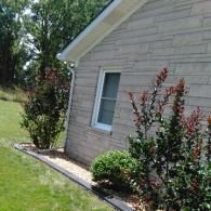 Side of a beige house with a window, shrubs, and green lawn in bright sunlight