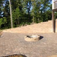 Stone patio with circular fire pit in front of a beige retaining wall and trees.