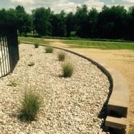 Curved landscaped gravel bed with small shrubs beside a concrete path and grass field under trees