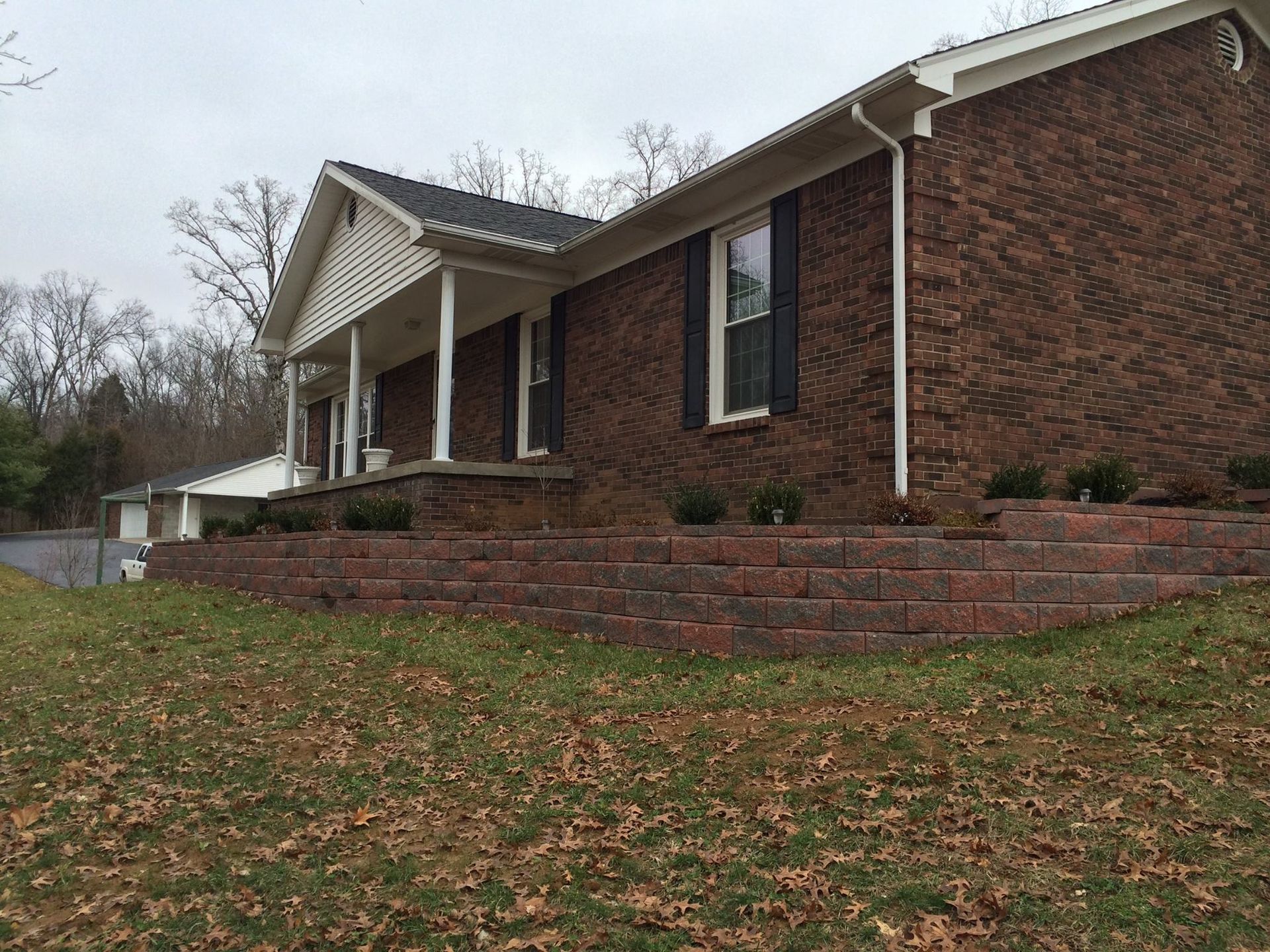 Brick house with front porch and stone retaining wall on a grassy, leaf-covered slope