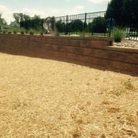Mulched yard with a brick retaining wall and black metal fence beside trees and shrubs