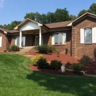 Brick suburban house with a front porch, red steps, and a green lawn in bright sunlight