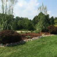 Landscaped garden with tall ornamental grasses, shrubs, rocks, and a green lawn under a blue sky