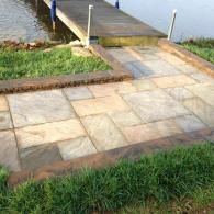 Stone walkway leading to a small dock over water, bordered by grass and a brick edge.