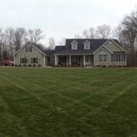 Large gray house with a porch and dormer windows behind a wide grassy lawn under a cloudy sky