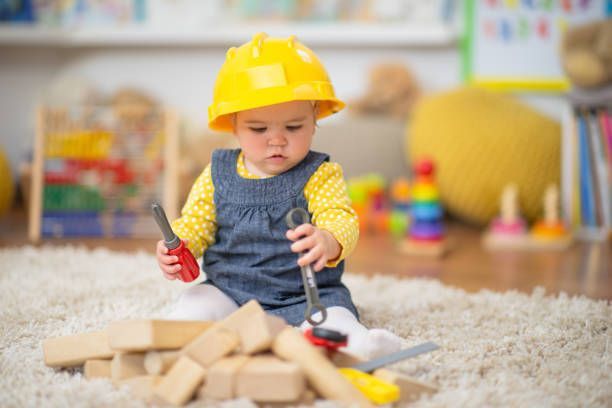A baby wearing a hard hat is playing with wooden blocks and a screwdriver.