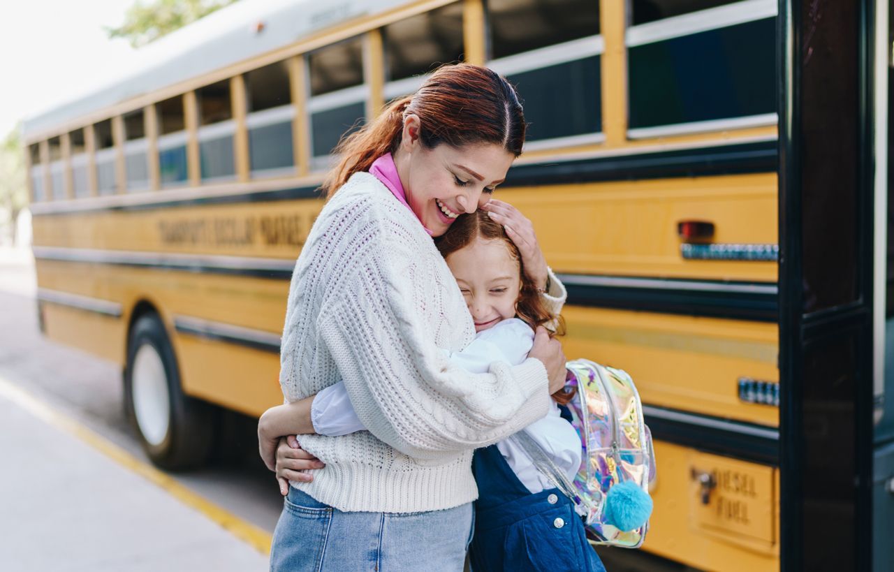 A woman is hugging a little girl in front of a school bus.