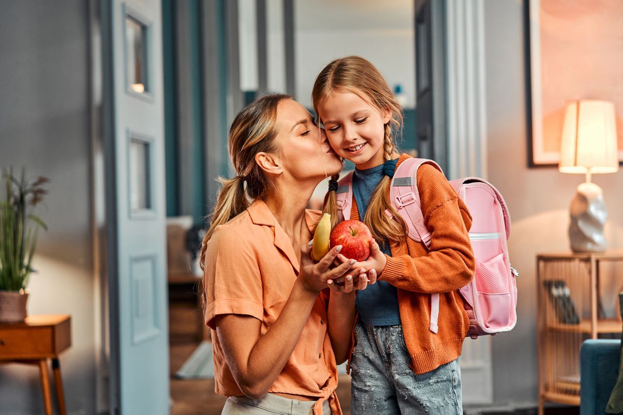 A woman is kissing a little girl on the cheek while the girl is holding an apple and a banana.