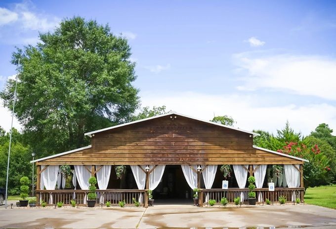 Wooden barn with white curtains, a porch, and a large tree against a blue sky.