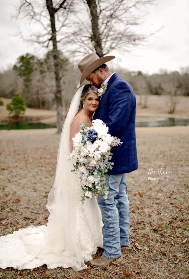 Bride and groom embrace in a field; man in cowboy hat and blue blazer kisses bride's head, holding bouquet.