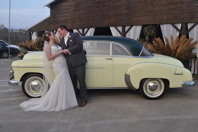 Newlyweds pose by a vintage yellow car in front of a barn-style building.