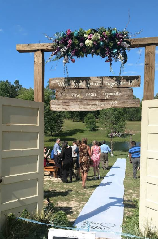 Wedding ceremony setup with floral arch, rustic wooden sign, and guests walking down the aisle.