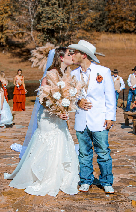 Bride and groom kissing outdoors; she in white gown and veil, he in white jacket, jeans, and cowboy hat.