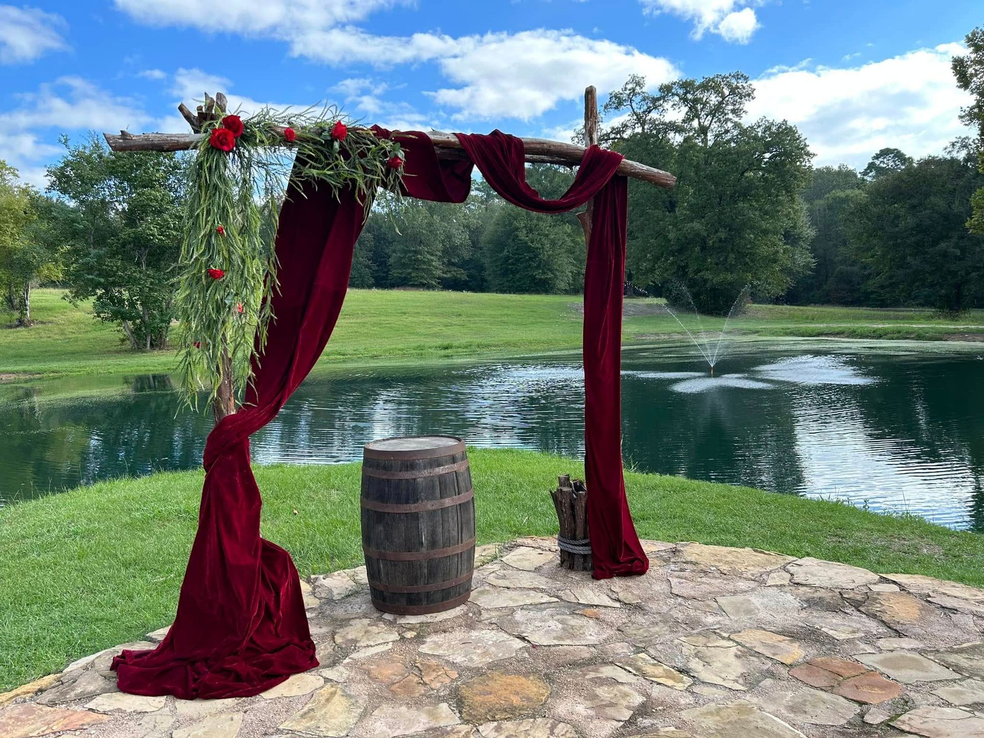 Wedding arch draped with red fabric and flowers overlooking a pond. Barrel and stone patio.