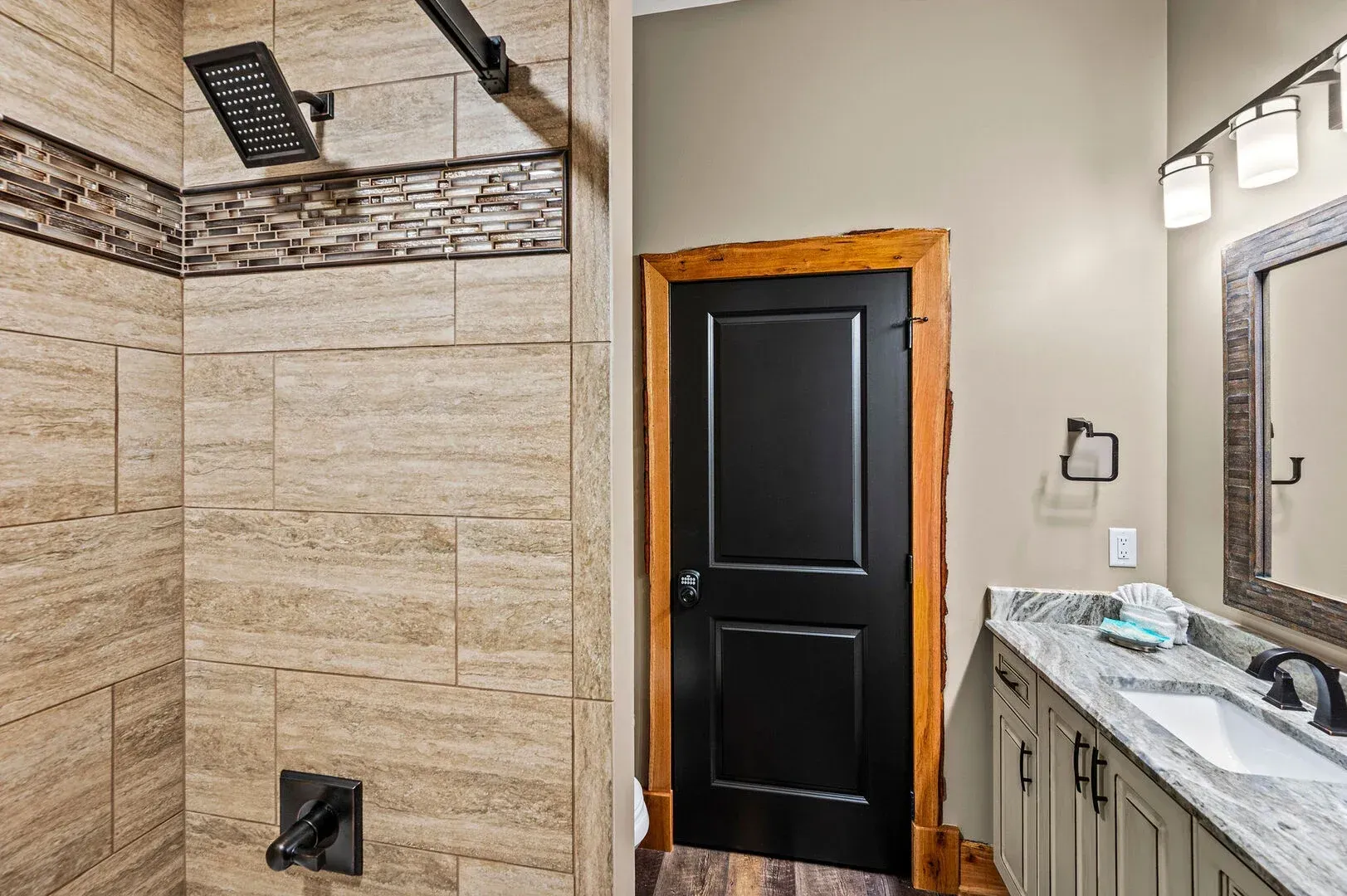 Bathroom with tiled shower, black door, wooden trim, gray vanity, and mirror.