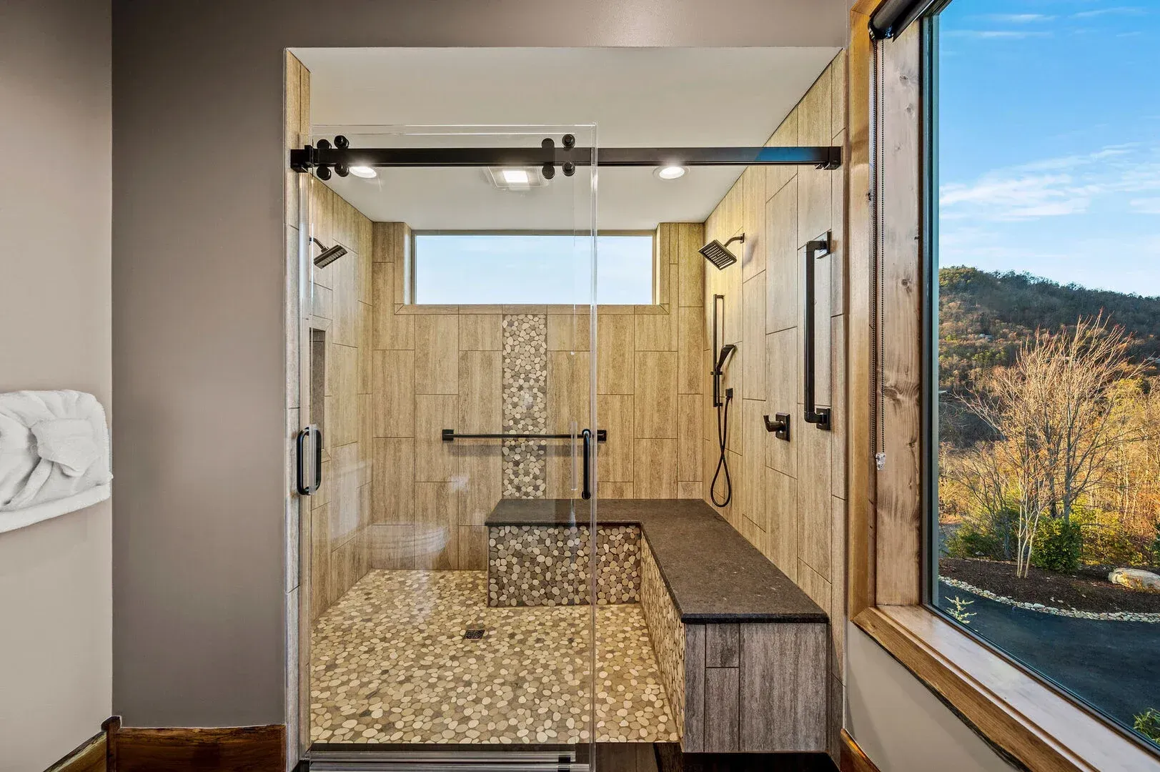 Modern shower with glass doors, stone tile, built-in bench, and a mountain view through a large window.