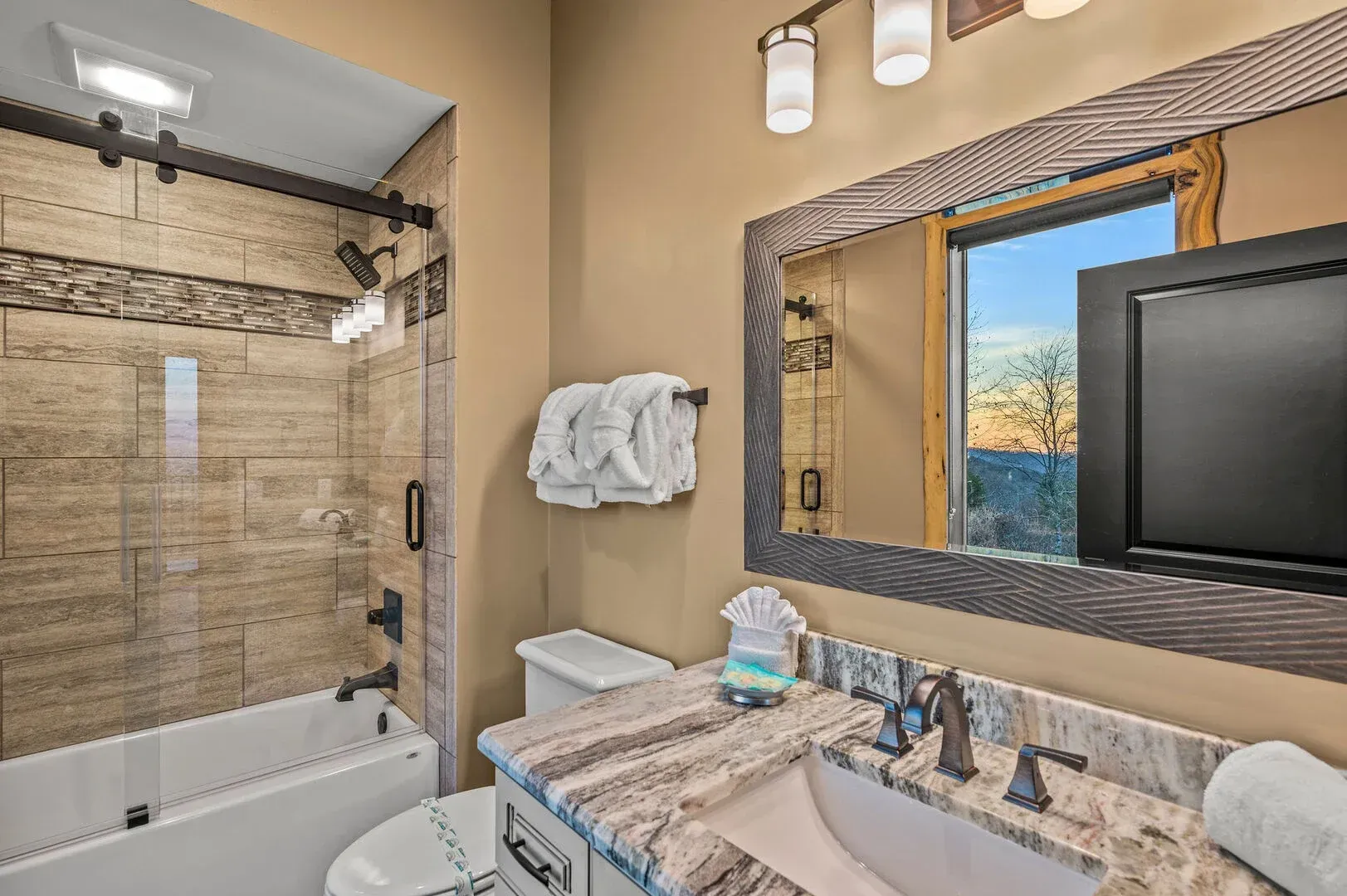 Bathroom with shower, toilet, vanity, large mirror, and TV; beige walls, granite countertop.