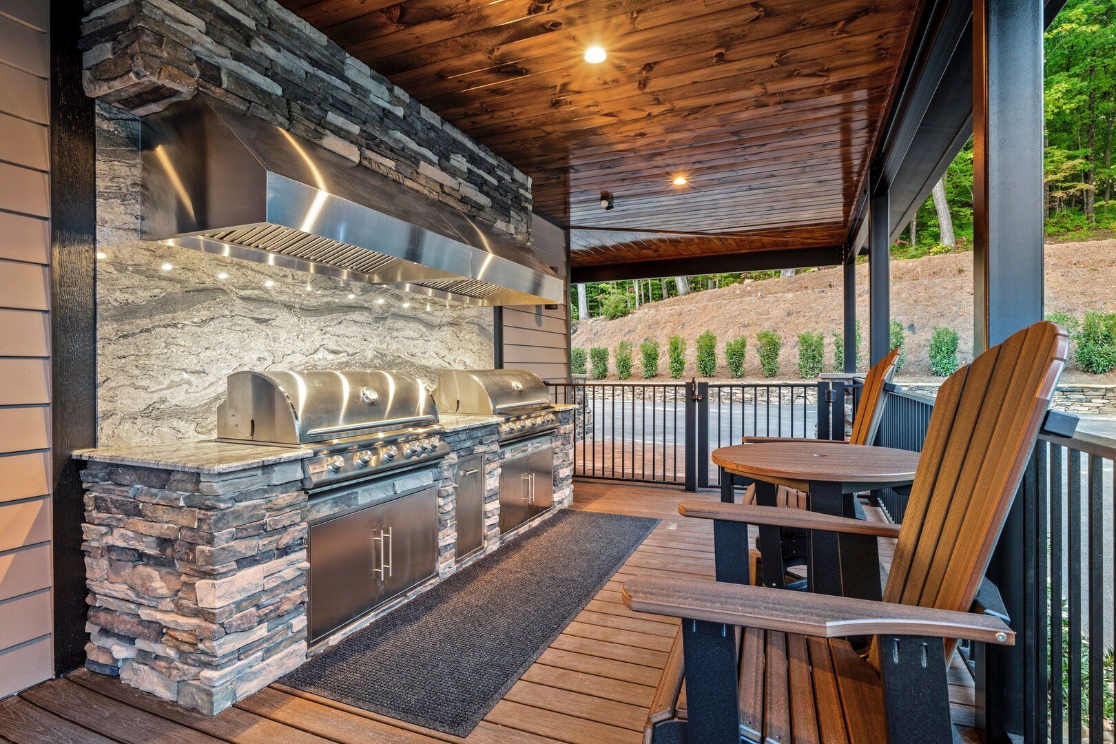 Outdoor kitchen with stone accents, stainless steel grill, wooden ceiling, and Adirondack chairs.
