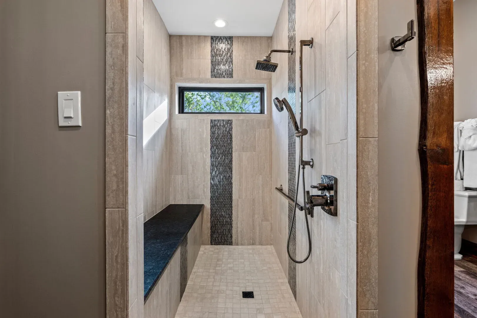 Walk-in shower with bench and rainfall shower head. Light beige tile, dark accents, and window.