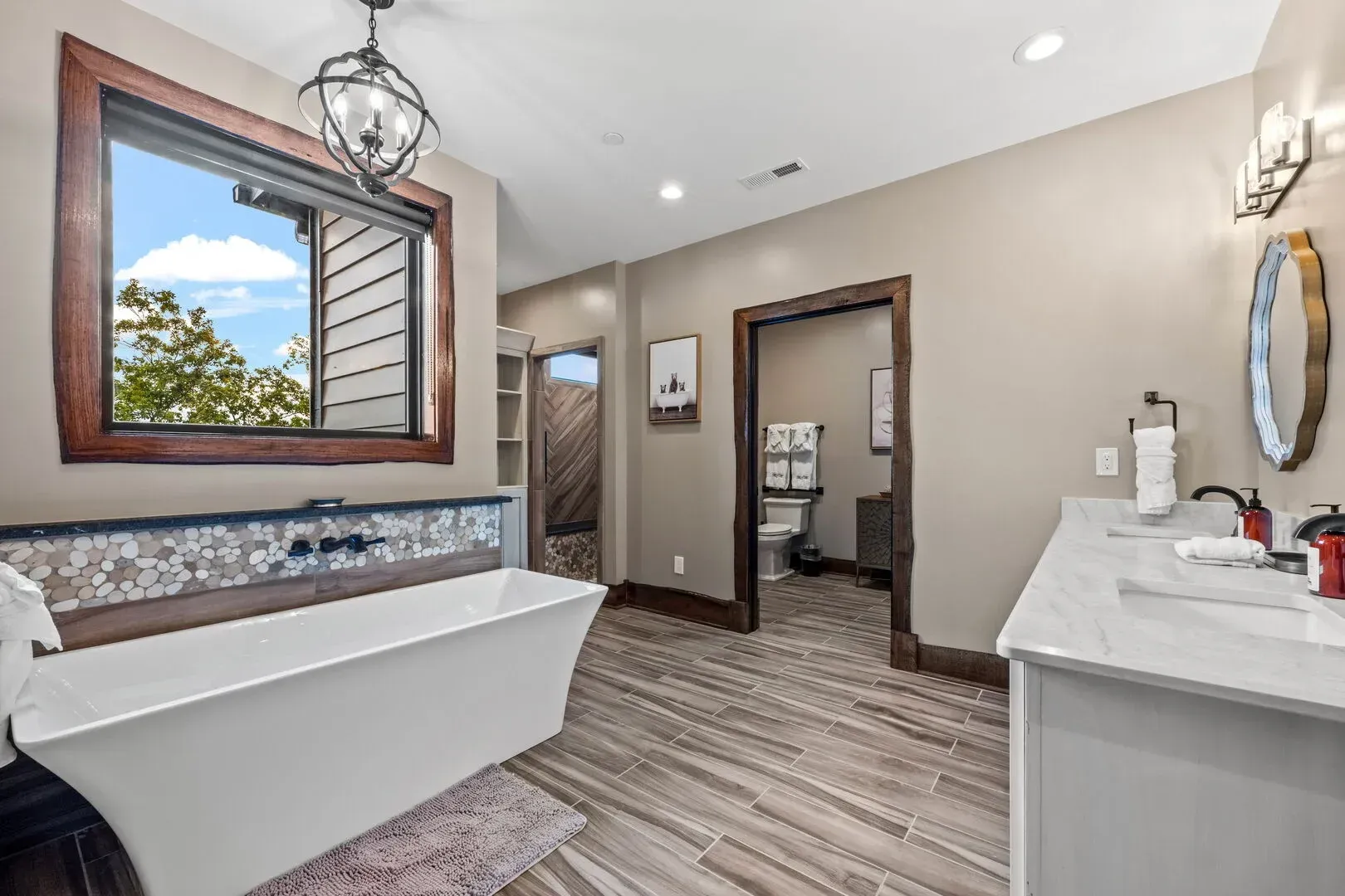 Bathroom with a white tub, double vanity, wood trim, and a window overlooking trees.