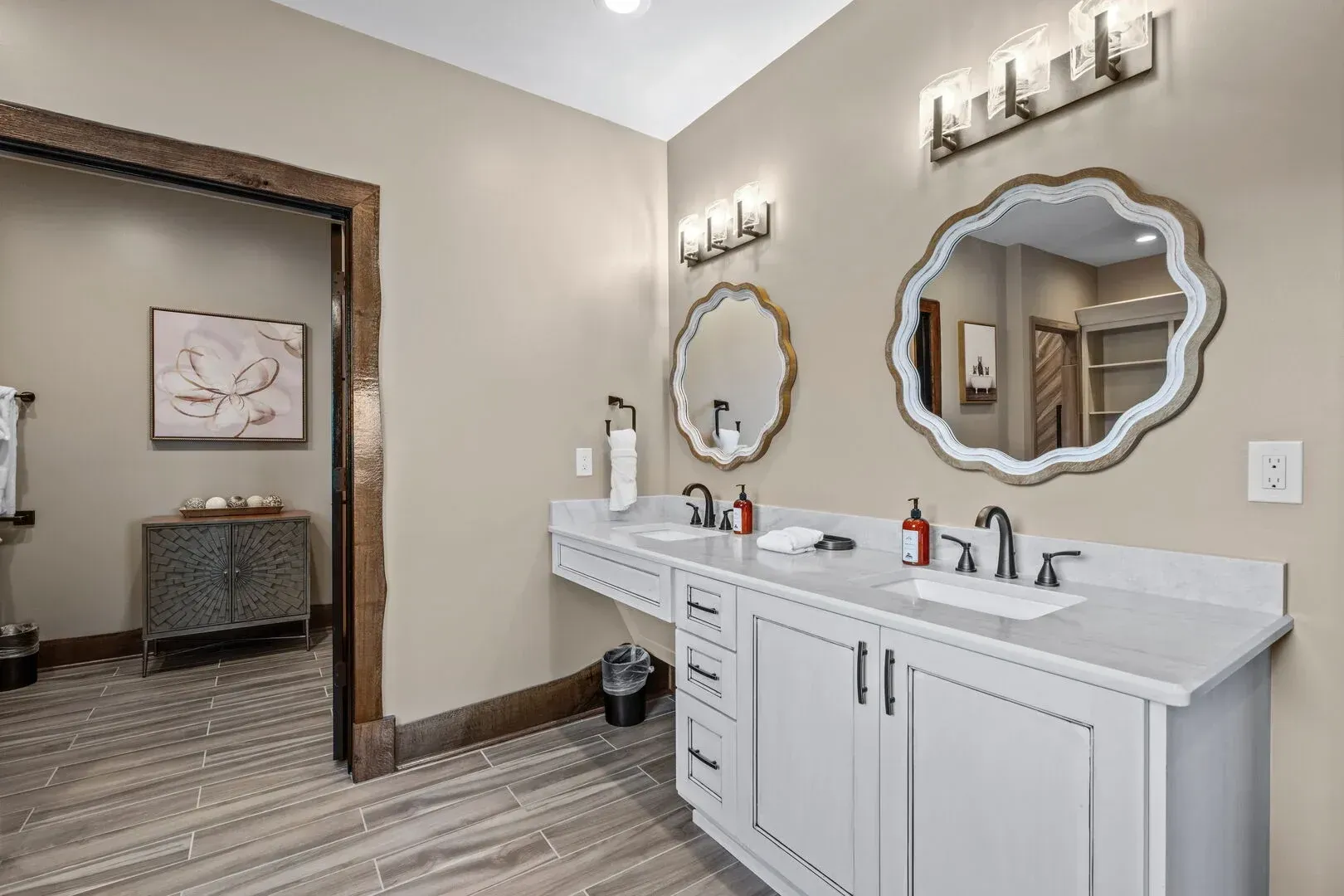 Modern bathroom with double sinks, mirrors, and beige walls. Open doorway leads to a sitting area.