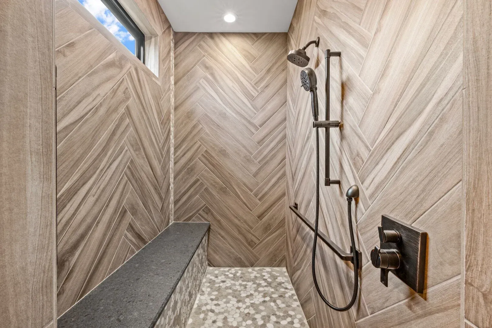 Shower with herringbone tile walls, built-in bench, pebble floor, and dark bronze shower fixtures.