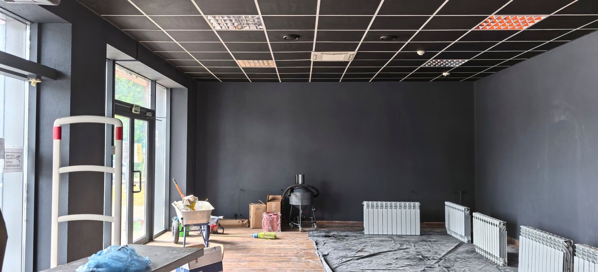 Empty room under renovation with dark walls, exposed ceiling grid, and tools on a worktable
