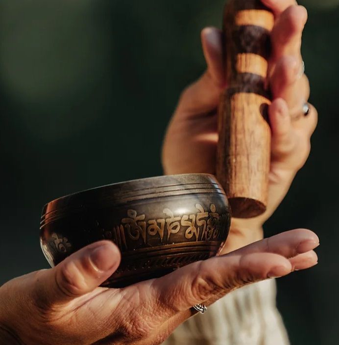 Handen die een bronzen klankschaal en een houten klopper vasthouden, buiten, wazige achtergrond.