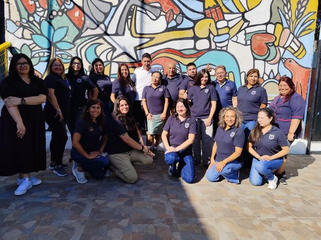 Grupo de personas posando frente a un colorido mural; muchos visten polos azules y pantalones vaqueros.