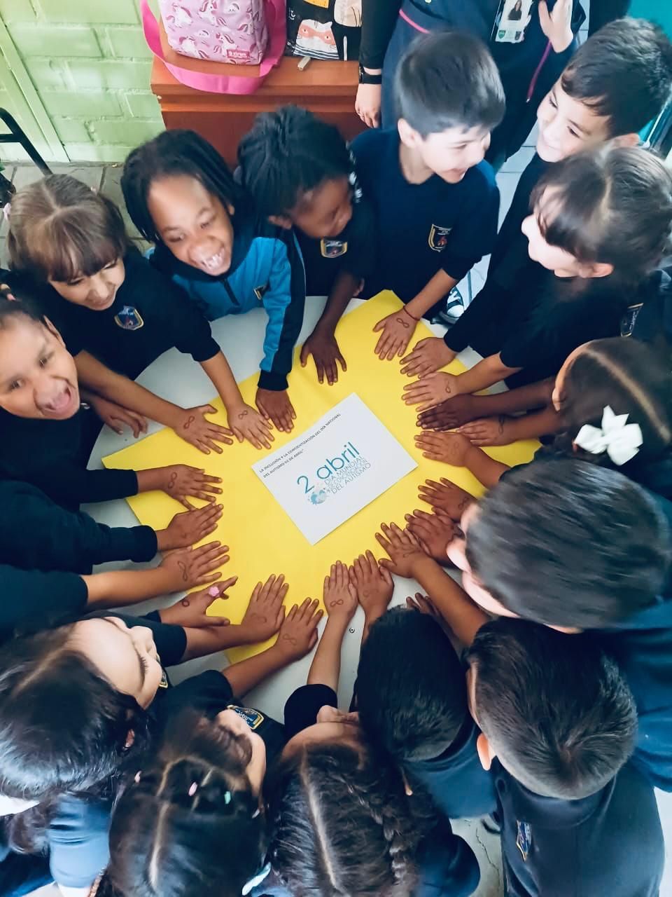 Niños alrededor de una figura amarilla con las manos sobre ella, sonriendo en un aula.