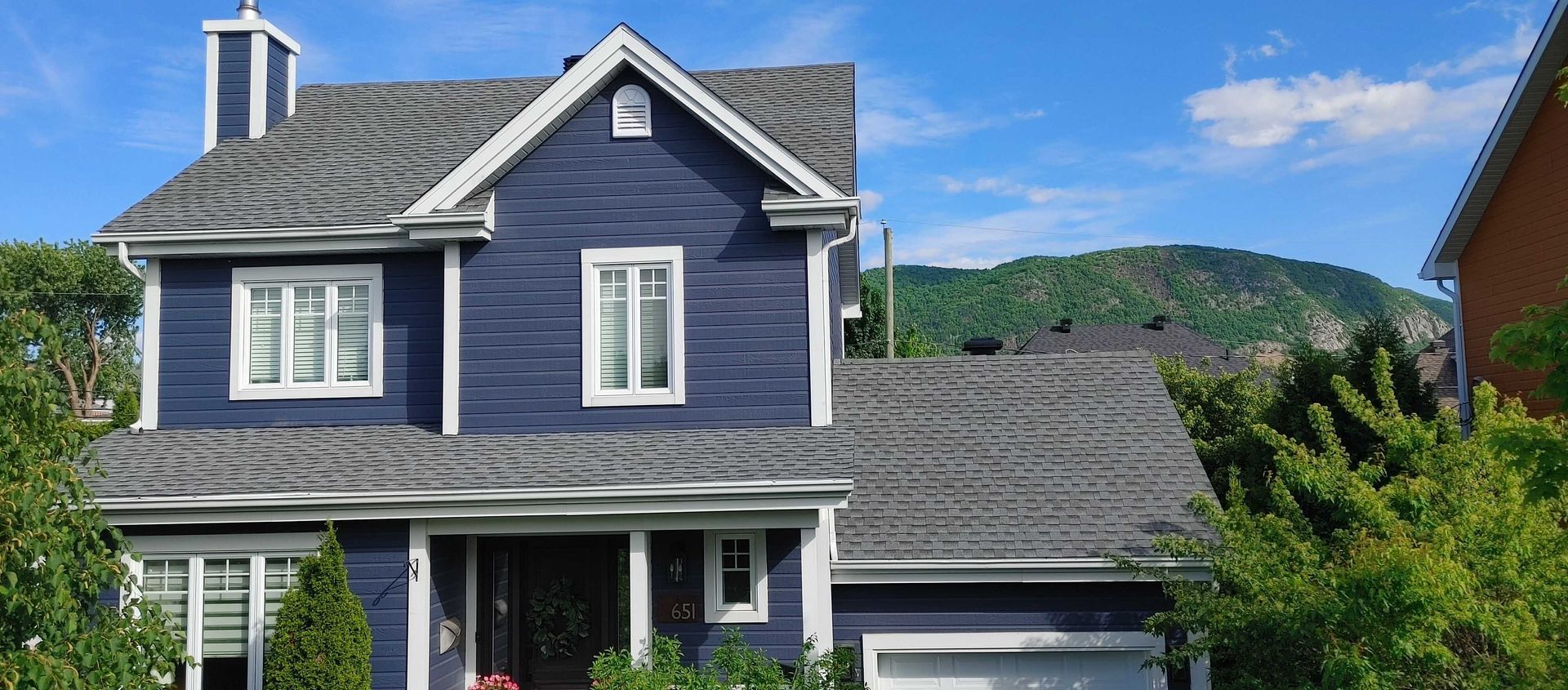 Maison bleue au toit gris et aux boiseries blanches. Arbres devant, colline verte en arrière-plan. Journée ensoleillée.