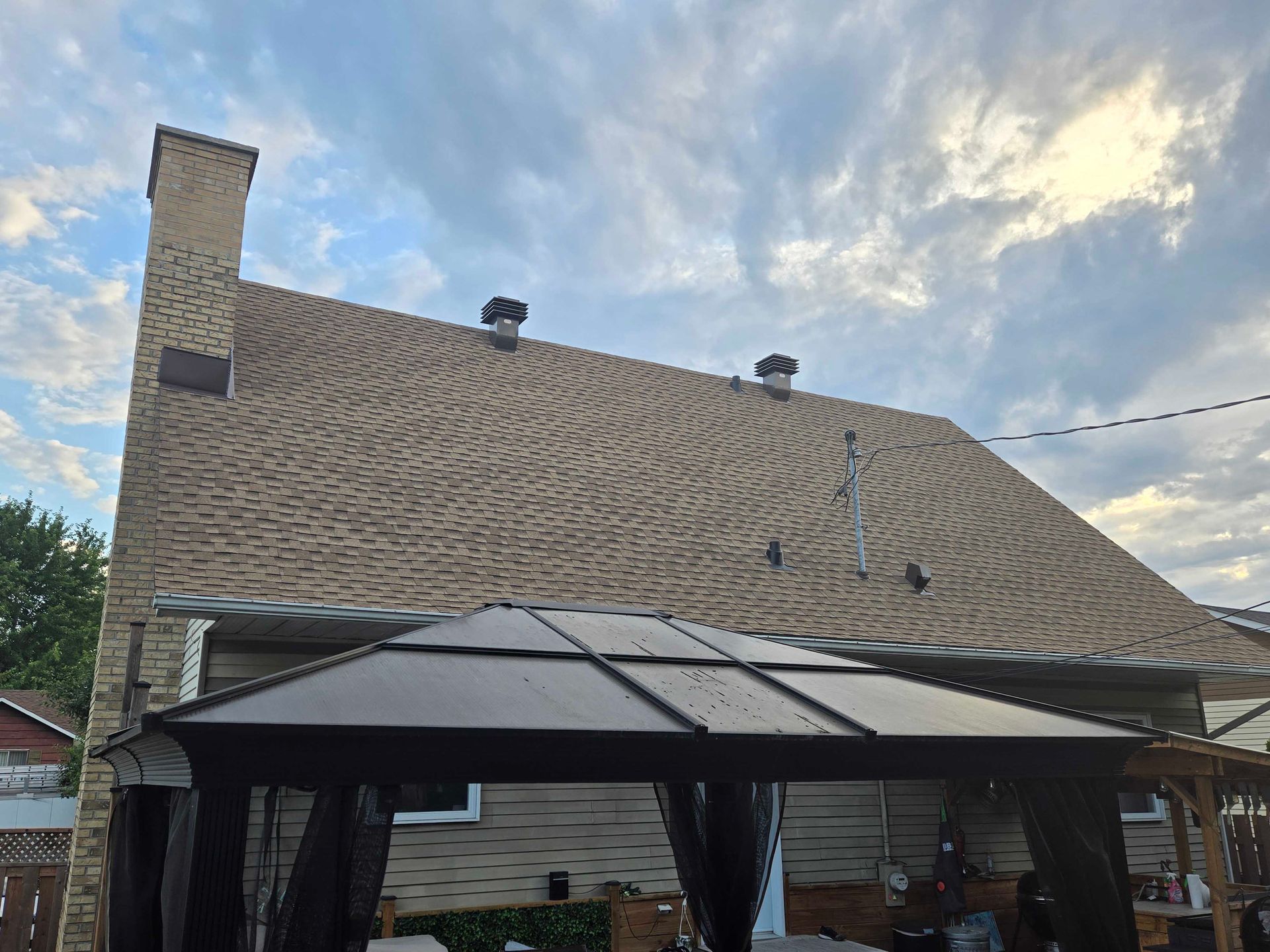 Toiture en bardeaux beiges avec une cheminée en briques et deux aérations noires, vue depuis une cour arrière sous un kiosque. Ciel nuageux.