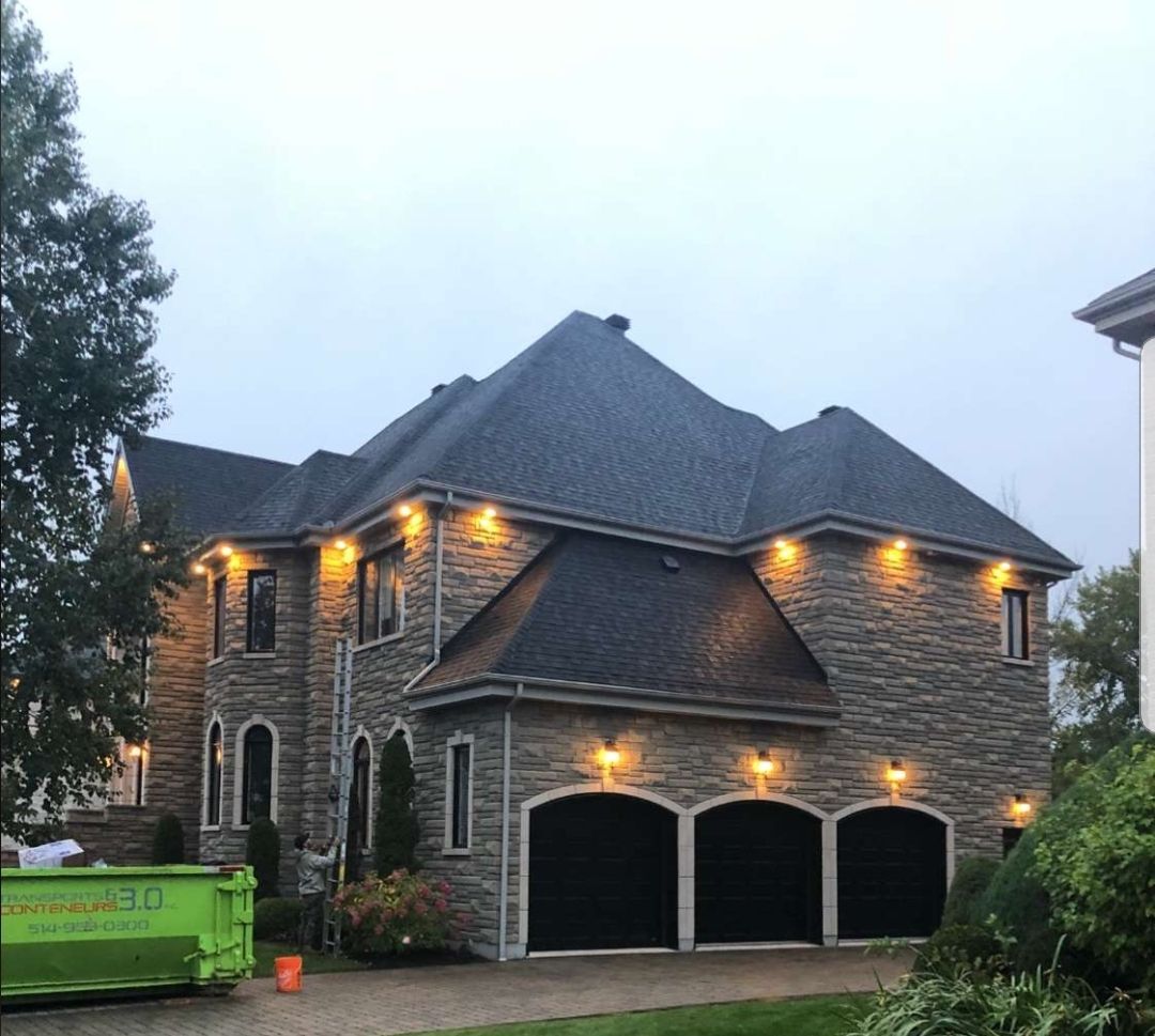 Maison revêtue de pierre, toit noir, garage pour trois voitures et éclairage extérieur chaleureux. On aperçoit une benne à ordures verte.